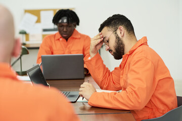 Young adult man sitting at desk holding head in hand studying with laptop, while young adult Black...