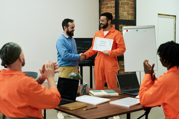 Middle aged man congratulating young adult inmate in orange prison uniform receiving certificate,...