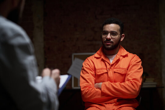 Young adult man sitting with arms crossed wearing prison uniform engaging in group therapy session with unseen counselor holding clipboard in correctional facility setting