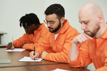 Three adult men sitting at table writing on paper during educational class in prison setting,...