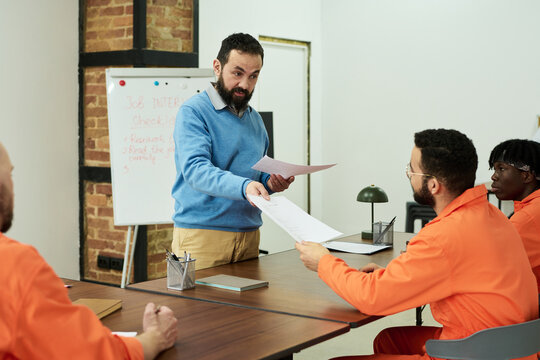 Middle aged Caucasian man handing documents to seated group of young adult men wearing orange uniforms, participating in classroom learning session in prison