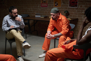 Caucasian man in orange prison uniform sitting with head down during group therapy session, with...