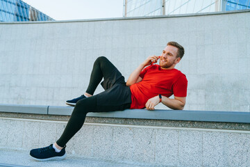 A man in a red shirt and black leggings relaxes on a concrete bench, engaging in a cheerful phone conversation under a clear sky.
