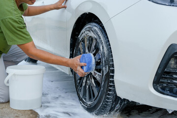 A smiling man is washing his car with soapy water under bright sunlight, showing his enthusiasm and joy in his daily work.