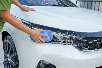 A smiling man is washing his car with soapy water under bright sunlight, showing his enthusiasm and joy in his daily work.
