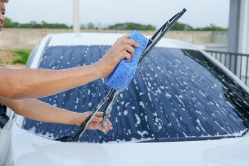 A smiling man is washing his car with soapy water under bright sunlight, showing his enthusiasm and joy in his daily work.