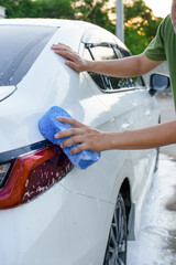 A smiling man is washing his car with soapy water under bright sunlight, showing his enthusiasm and joy in his daily work.