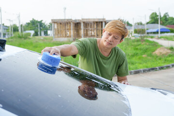 A smiling man is washing his car with soapy water under bright sunlight, showing his enthusiasm and joy in his daily work.