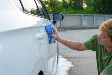 A smiling man is washing his car with soapy water under bright sunlight, showing his enthusiasm and joy in his daily work.