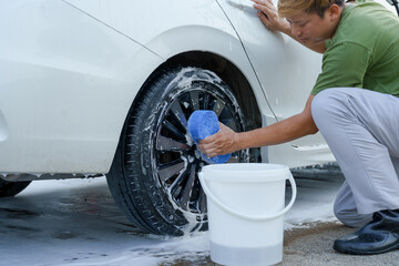 A smiling man is washing his car with soapy water under bright sunlight, showing his enthusiasm and joy in his daily work.