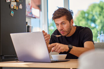 Man eating porridge while looking surprised at his laptop screen during breakfast. Person has...