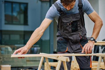 Man measuring wooden board during carpentry work in front of residential building. Concept of...