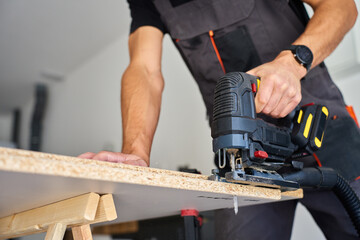 Carpenter cuts wooden board with electric jigsaw in home workshop. Man in workwear working with...