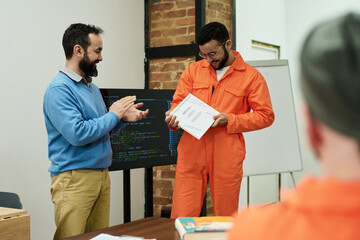 Middle aged man applauding young adult man wearing prison uniform receiving certificate in...
