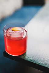 Refreshing red cocktail with orange peel garnish and large ice cube in a clear glass on wooden table, close-up with blurred background.