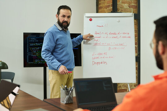 Middle aged Caucasian man teaching programming concepts on whiteboard to adult male student in classroom setting, discussing code structure with laptops and digital screen visible - Powered by Adobe
