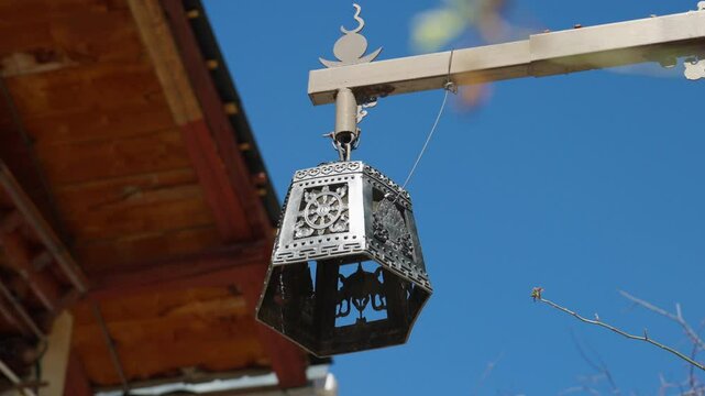An intricately patterned lantern hangs from a beam against a blue sky&mdash;highlighting cultural design, crafted textures, and a peaceful blend of tradition and nature.