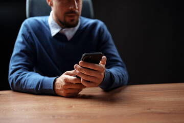 Man in a blue sweater using smartphone while sitting at a wooden desk in a dimly lit room