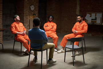 Group of young adult male convicts sitting in circle wearing prison uniforms participating in group...