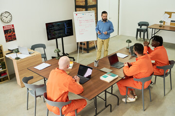 Caucasian middle aged man teaching coding to group of adult men in orange prison uniforms, diverse...
