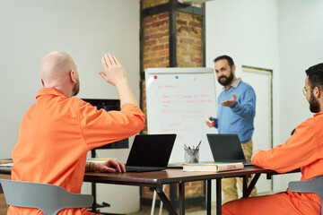 Middle aged Caucasian man teaching diverse group of adult men in orange prison uniforms using laptops, one adult man raising hand while sitting at table in classroom setting