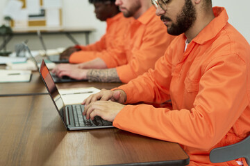 Group of young adult men of diverse ethnicities wearing orange uniforms sitting at desks using...