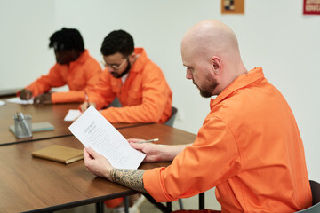 Caucasian man with tattooed arm reading document at table while two inmates writing in background,...