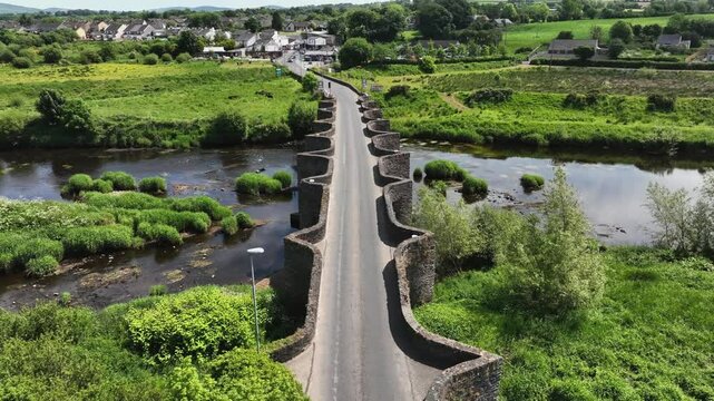 Clady Bridge, County Tyrone, Northern Ireland, June 2023. Drone pullback tilting up as car crosses over famed arch bridge with scalloped edge walls crossing River Finn.