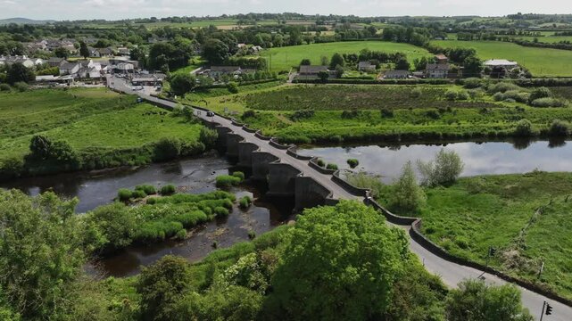 Clady Bridge, County Tyrone, Northern Ireland, June 2023. Drone medium orbit counterclockwise above famous bridge as cars cross over River Finn at midday with cloudy sky reflecting in water.