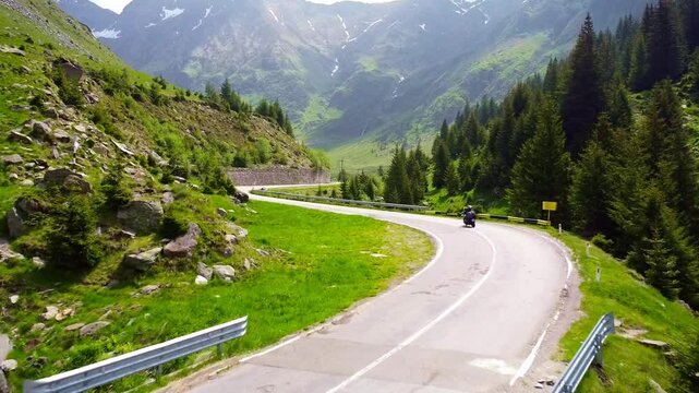 Drone tracks biker riding through Transfăgărășan road in Carpathian Mountains of Romania, surrounded by lush greenery and alpine landscape.