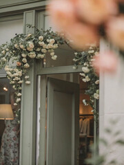 A sage green storefront with cascading cream roses, warm lighting, and a floral dress inside, creating a cozy, vintage-inspired vibe.