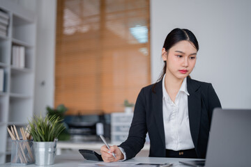 Young businesswoman wearing formal clothes sitting at desk working with laptop and taking notes using pen and calculator in modern office with plants and bookcase in background