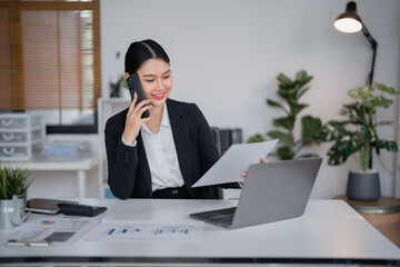 Asian businesswoman sitting at a modern desk, engaged in a phone conversation while analyzing documents and working efficiently on a laptop surrounded by vibrant office plants