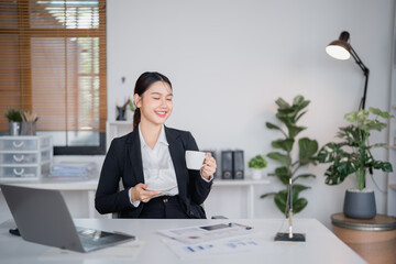 Young asian businesswoman in suit drinking coffee during break at workplace, sitting at desk with...