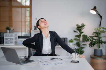 Young asian businesswoman having back pain while sitting on chair in office, suffering from backache after long working on laptop computer, office syndrome concept