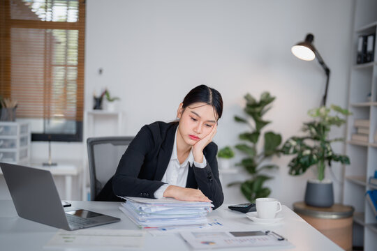 Asian businesswoman resting head on hand and looking down at stack of paperwork on desk while working with laptop computer in office, feeling tired, bored, and stressed from work