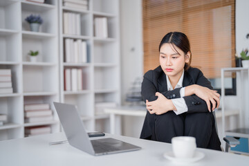 Asian businesswoman feeling stressed and exhausted at modern office, struggling with headache and burnout while working on laptop against looming deadlines