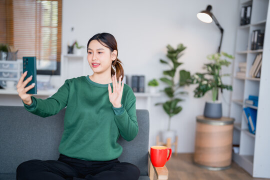 Asian freelancer woman wearing green sweater sitting on comfortable sofa waving and smiling during video call with smartphone in cozy home office