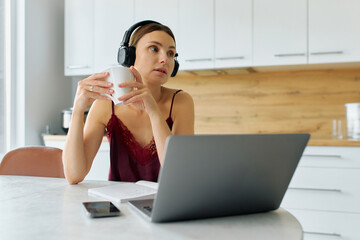 Young woman dressed in pajamas drinking coffee and having video meeting using laptop and headphones in kitchen