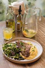 Plate of grilled steak served with creamy mashed potatoes and mixed greens, accompanied by a glass of juice and a pitcher of infused water on a wooden table, showcasing a casual dining setup.