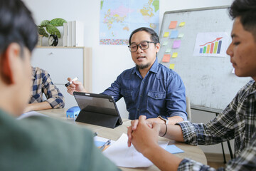 Teacher Wearing Glasses Holds Marker And Speaks While Looking At Tablet In Meeting With Students
