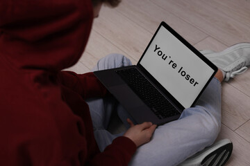 Cyber bullying. Upset teenage boy looking at laptop with text You Loser on floor indoors, closeup