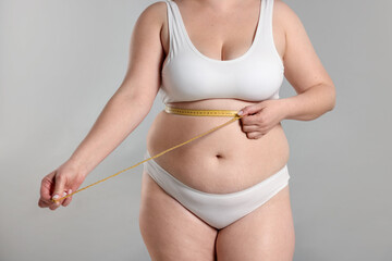Weight loss. Plus size woman measuring waist with tape on gray background, closeup