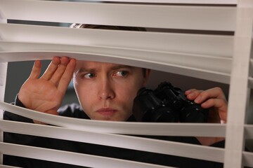 Paranoia. Worried man looking through window blinds indoors