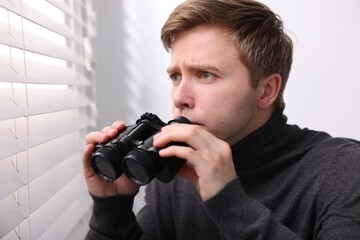 Paranoia. Worried man with binoculars near window indoors
