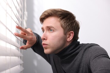 Paranoia. Worried man looking through window blinds indoors