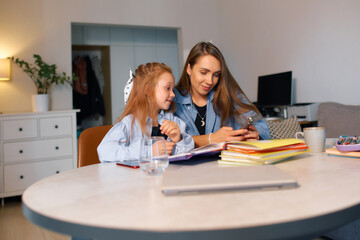 A young schoolgirl sits with her mother at the table and surfs the Internet before doing her...