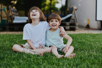 Fototapeta premium Two happy boys sitting on green grass, enjoying a sunny day while sharing laughter and friendship. A perfect moment of childhood joy captured in a natural setting.