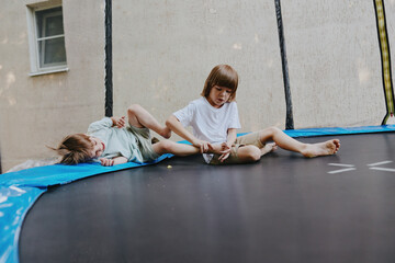 Two playful children, one sitting and the other lying down, enjoy a fun moment on a trampoline....