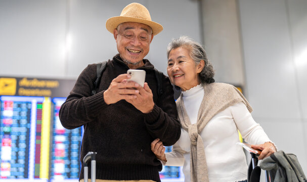Senior asian couple with luggage checking flight info on smartphone at airport, enjoying retirement travel, using travel booking app, digital technology, airline journey and senior trip planning